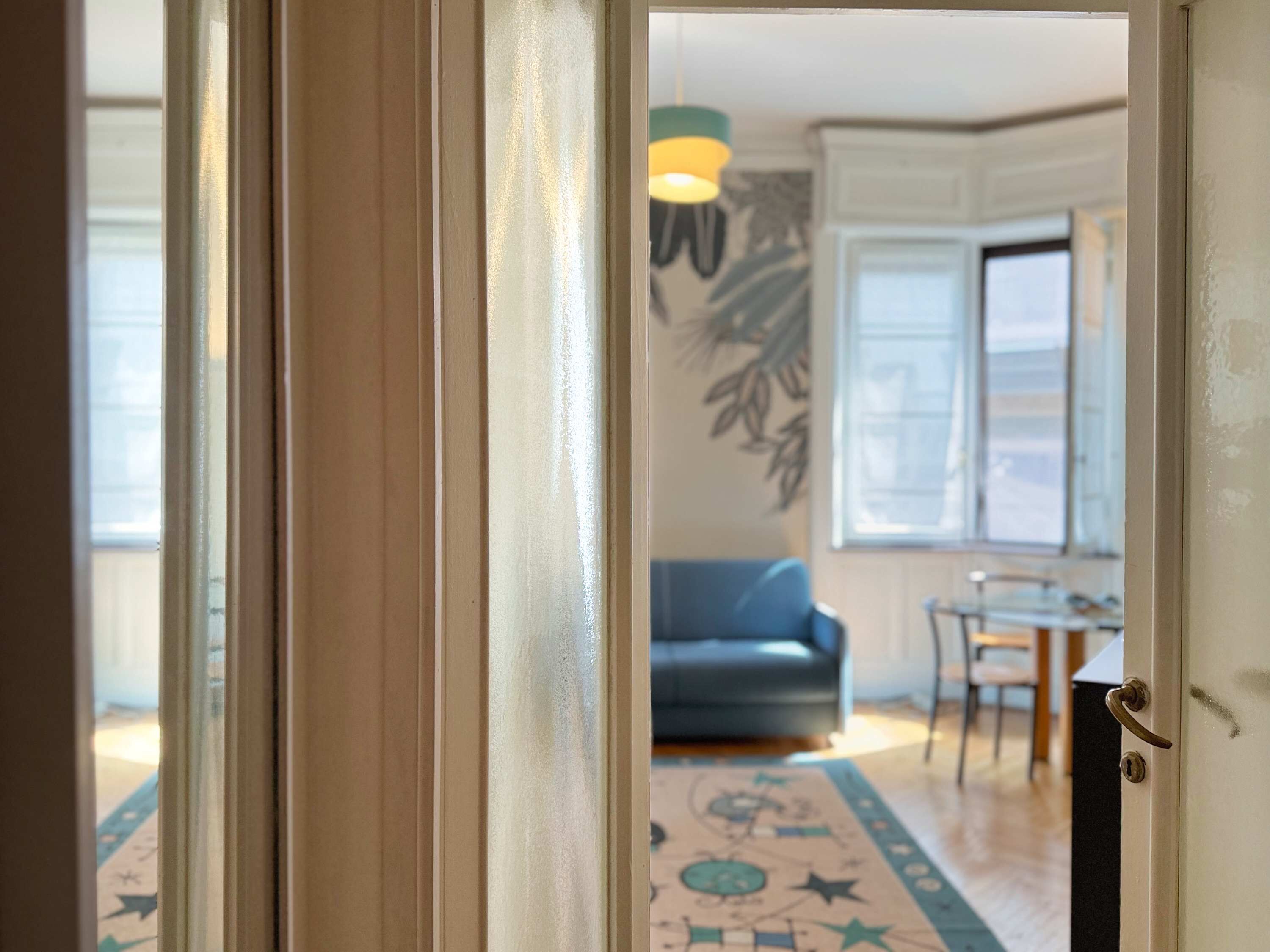 A room with a patterned rug, a blue sofa, a dining table, and a hanging light fixture.