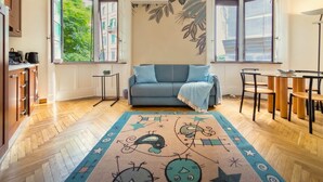 A room with a sofa, a dining table with chairs, a small table with a lamp, and a large window with a view of buildings and greenery. - Casa Nicole - fine stay in Navigli district (Milano)