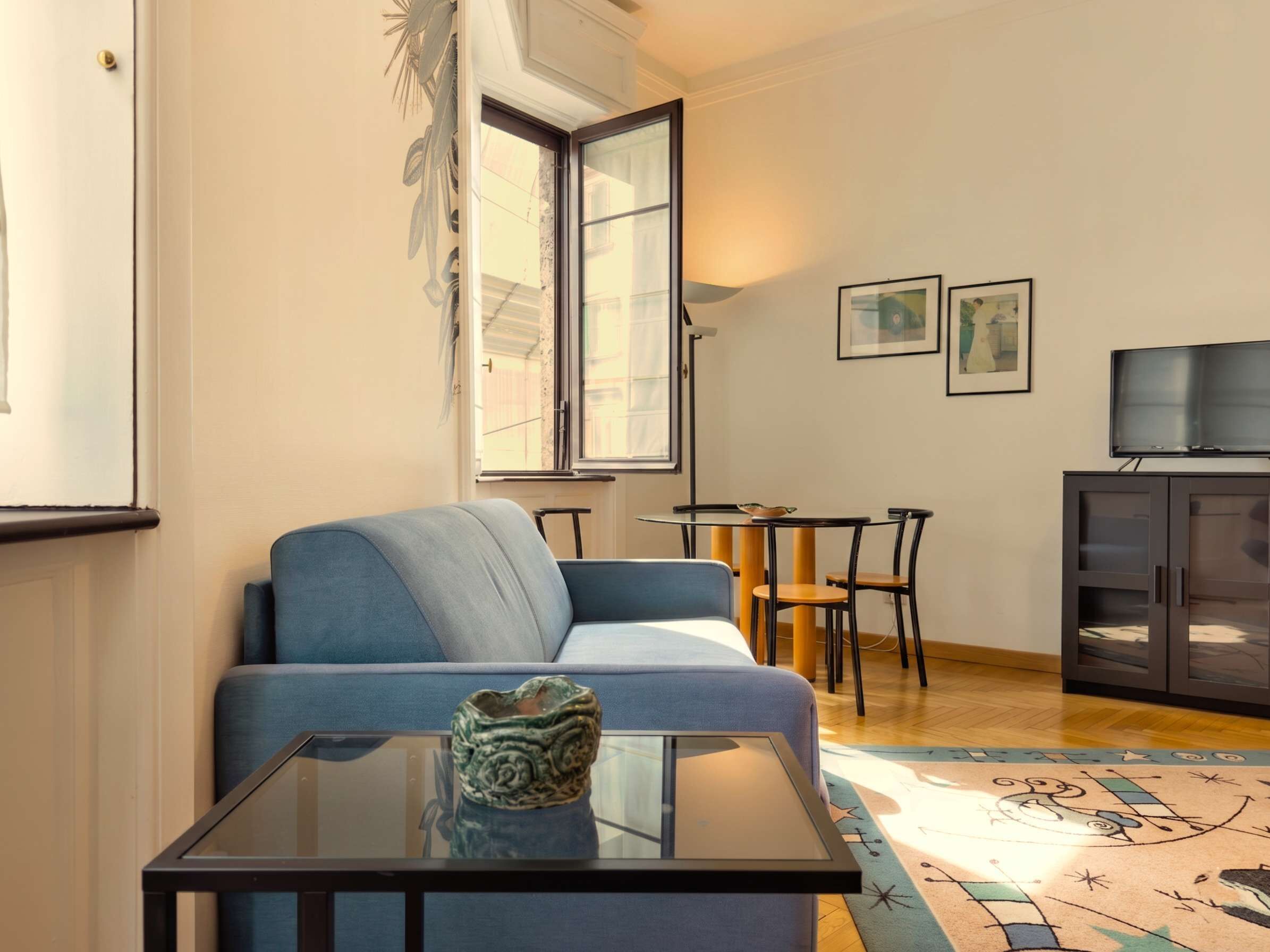 Un salon comprenant un canapé, une table basse en verre, une petite table avec des chaises, un téléviseur et des œuvres d’art accrochées au mur.