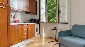 A compact kitchen with wooden cabinets, a washing machine, a microwave, and a window with a view of greenery. - Casa Nicole - fine stay in Navigli district (Milano)