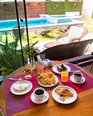 Una mesa de desayuno con diversos alimentos y bebidas, una piscina al fondo y mobiliario de exterior.