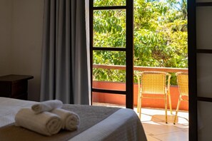 Habitación de hotel con cama, un par de toallas enrolladas, ventana con vistas a la vegetación y balcón con sillas de mimbre.