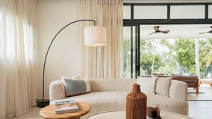 Een moderne woonkamer met een beige bank, een ronde salontafel, een vloerlamp en een groot raam met uitzicht op groen.