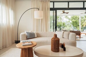 A modern living room with a beige sofa, a round coffee table, a floor lamp, and a large window with a view of greenery. - Modern 2BR Condo w/ Beach Views • Sol de Arena A6 (Las Terrenas)