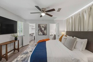 Ein Hotelzimmer mit Bett, Deckenventilator, Fernseher, Pflanze, Gemälde und angrenzendem Badezimmer.