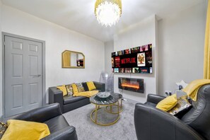 Un salon avec une porte grise, un canapé noir orné de coussins jaunes, une table basse en verre et un téléviseur fixé au mur qui affiche différents titres de films.