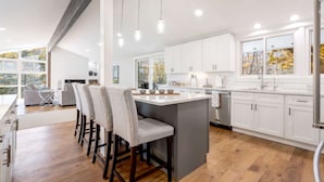A modern kitchen with white cabinets, a central island, bar stools, and pendant lighting. - Near Beach + Shopping • Tesla Charger • Large Deck (Kennebunkport)