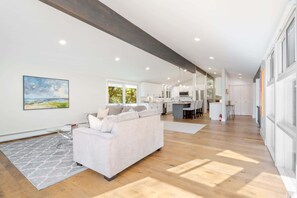 A spacious living area with a white sofa, a dining table, and a kitchen in the background.