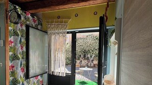 Een kamer met een tropisch gordijn, een macramé ophanging, een deur naar buiten en een kleine tafel met een stoel.