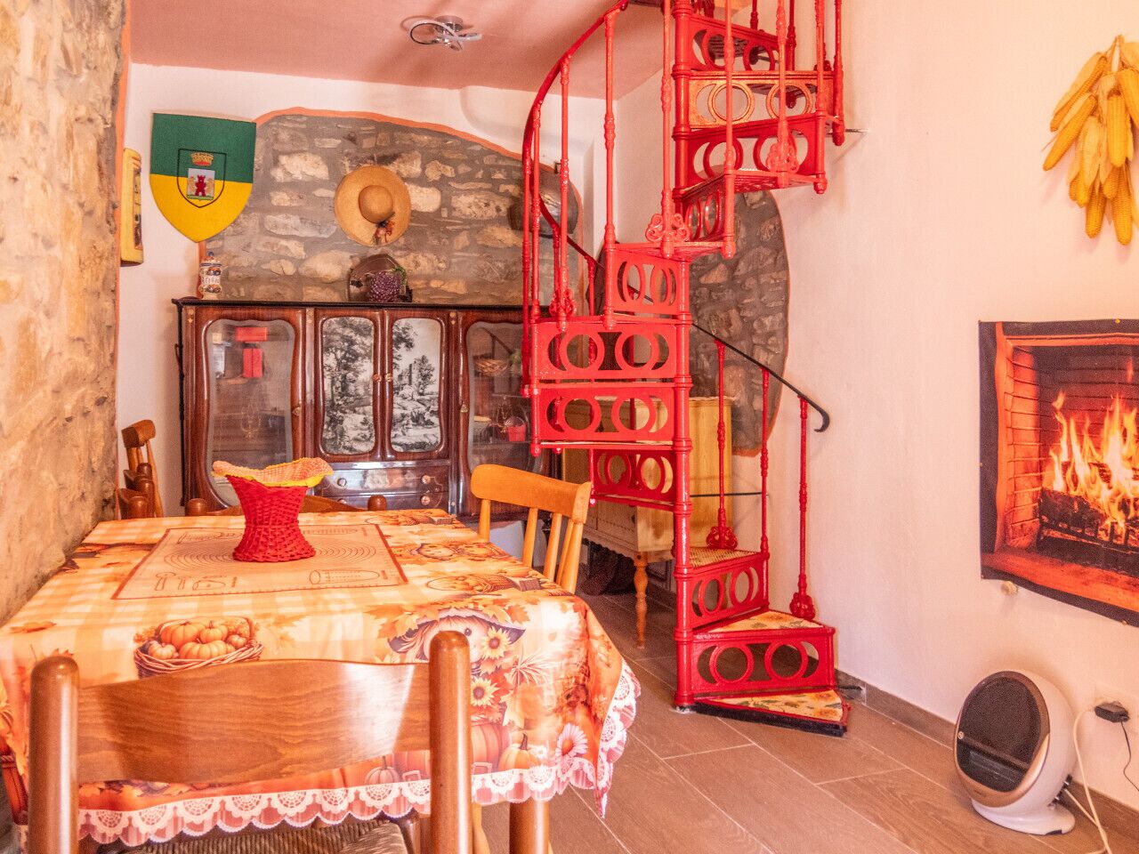 Un coin repas avec une table dressée pour deux personnes, un escalier en colimaçon rouge, un mur en pierre et une cheminée.