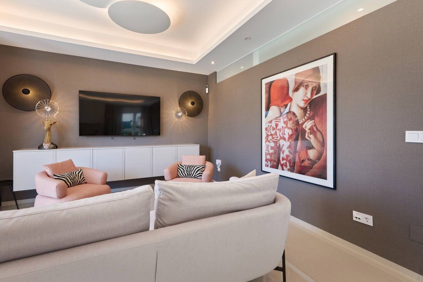 Un salon moderne comprenant un canapé, deux fauteuils, un téléviseur à écran plat, des appliques murales décoratives et un grand tableau encadré.