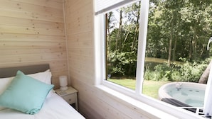 A wooden cabin room with a bed, a nightstand, a window, and a hot tub visible through the window. - Skylark (Ross-on-Wye)