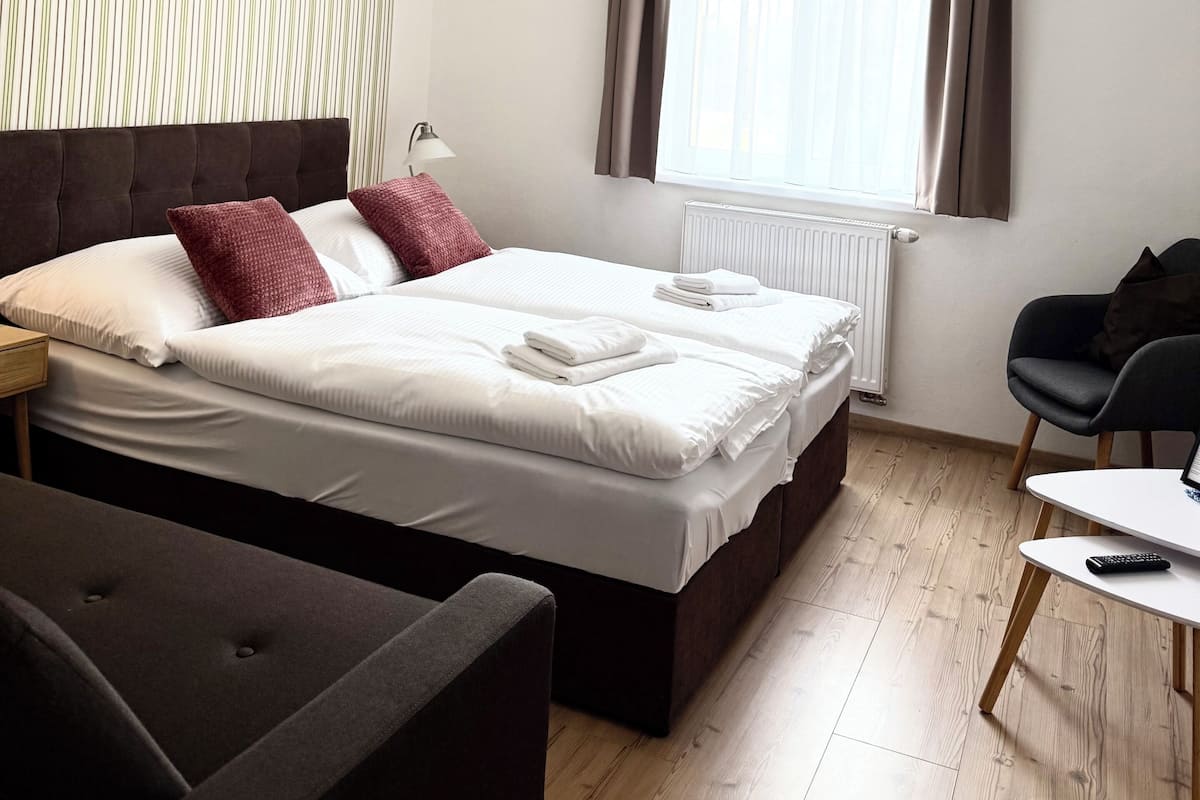 Habitación de hotel con cama, sillón, mesa, lámpara, ventana con cortinas y radiador.