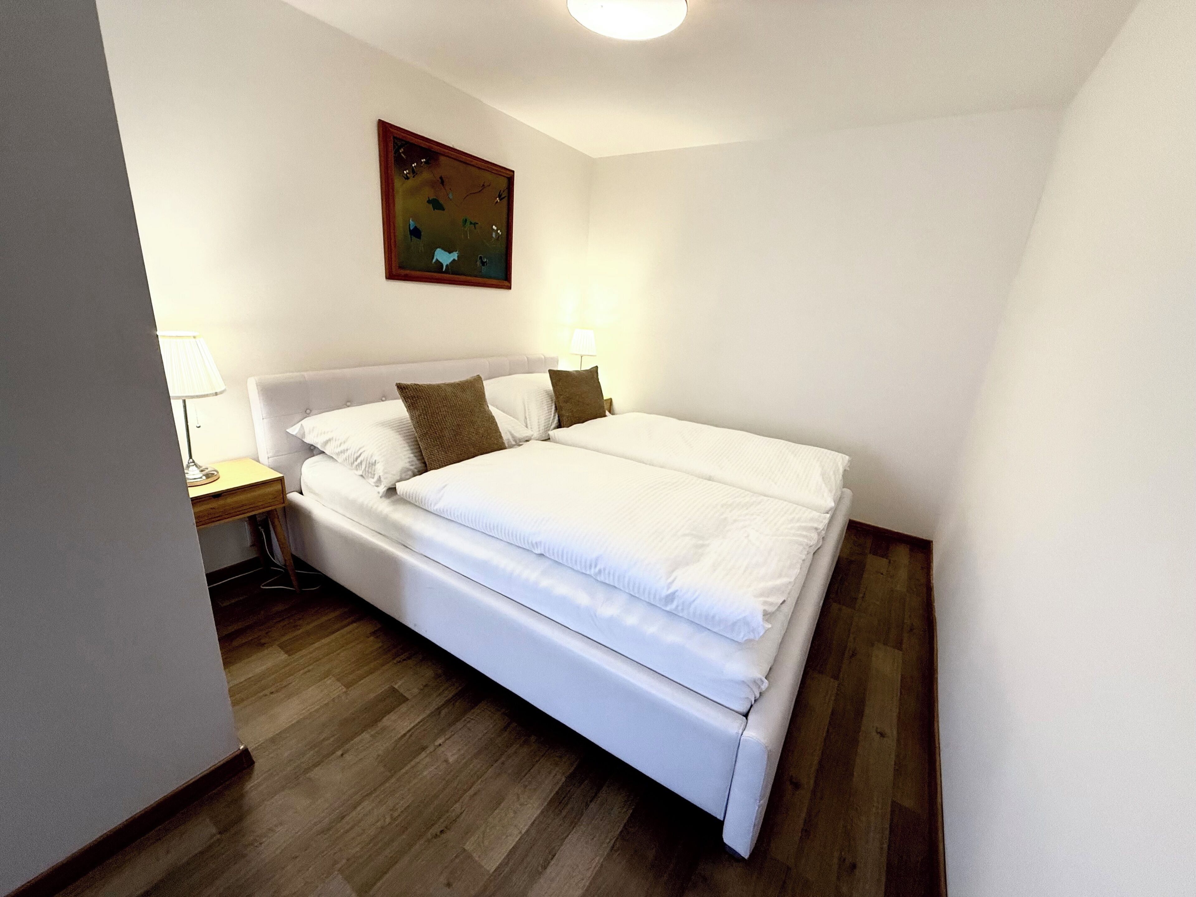 Un lit bien fait, avec du linge de lit blanc, une tête de lit en bois et une petite table de chevet surmontée d’une lampe.