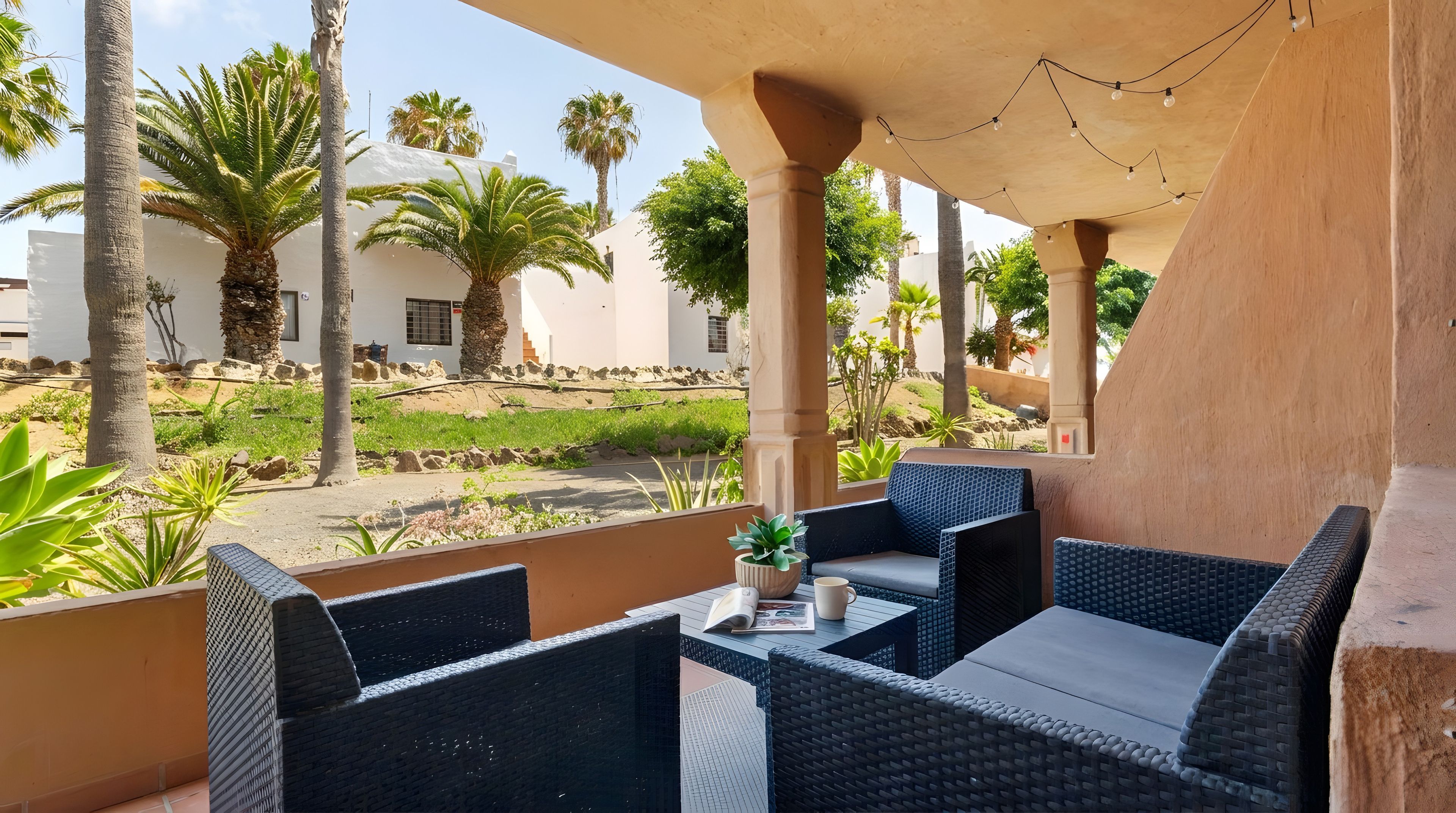 Un área de estar al aire libre con mobiliario de mimbre, una pequeña mesa con una planta y vistas a palmeras y edificios al fondo.