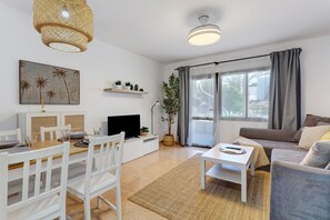 A modern living room with a dining area, a sofa, a TV, and a large window with curtains. - Oasis Duna _ Estrella Del Mar Pool with Shared Pool, Private Terrace, and Wi-Fi (Corralejo)