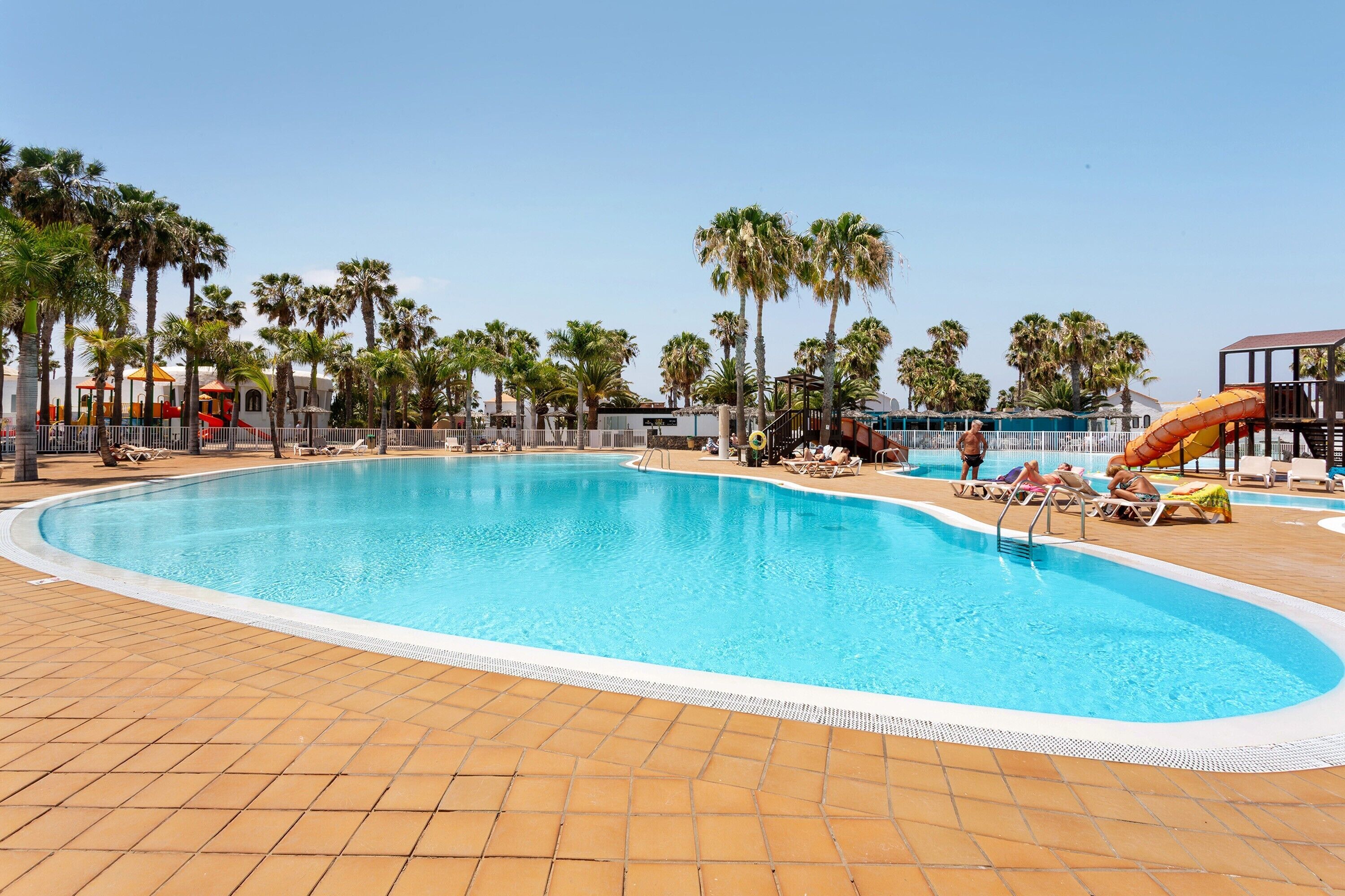 Ein Schwimmbecken mit gefliestem Sonnendeck, Palmen und einem Spielbereich für Kinder.