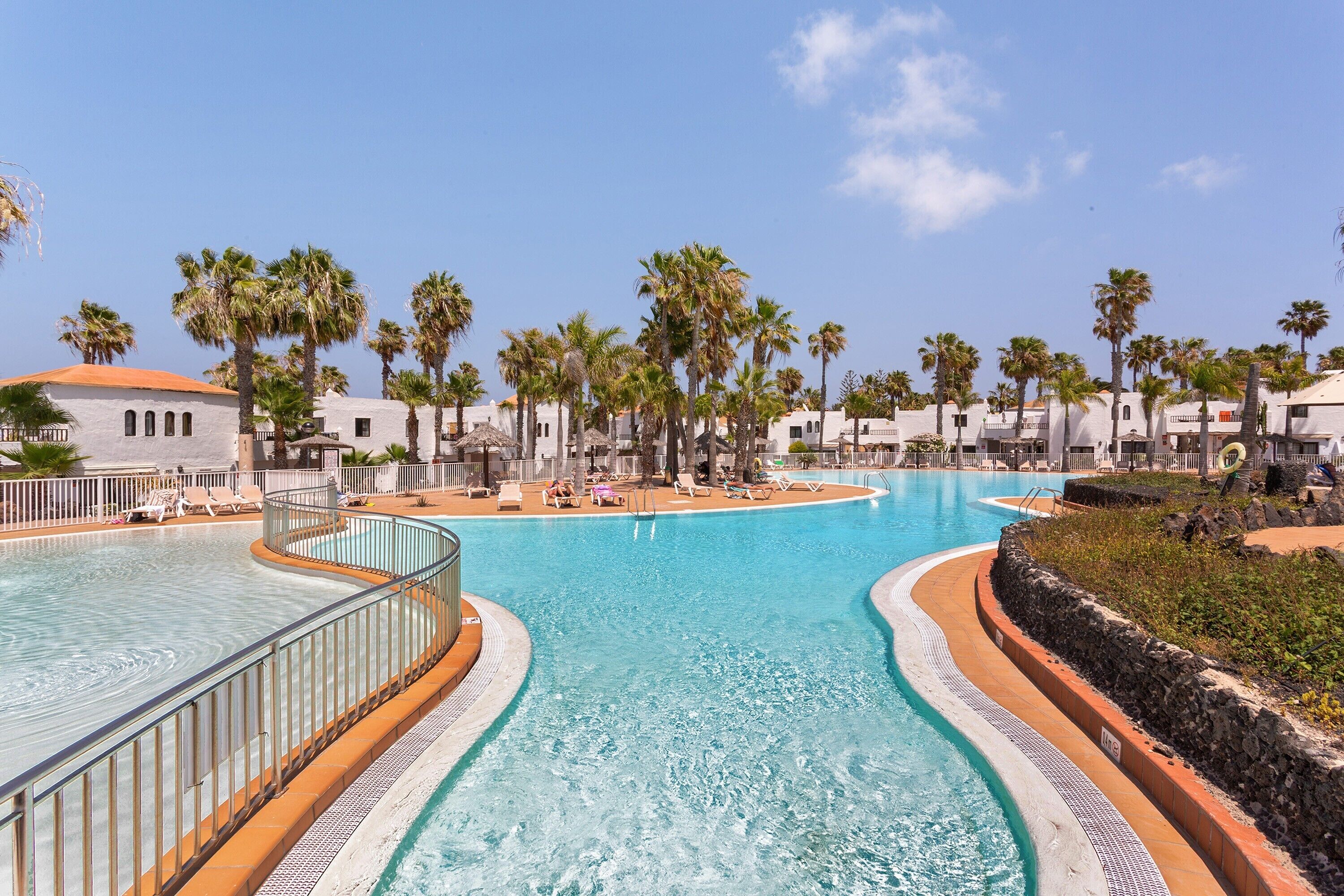 A large outdoor swimming pool with a curved design, surrounded by palm trees and white buildings.