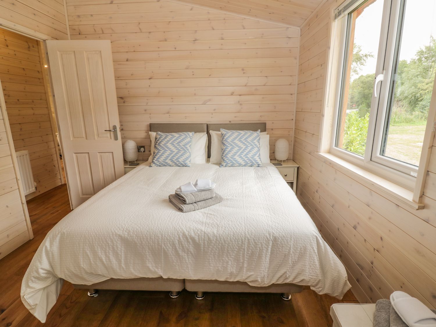 Un dormitorio con techo de madera, una cama grande con ropa de cama blanca, dos mesitas de noche y una ventana con vistas a la vegetación.