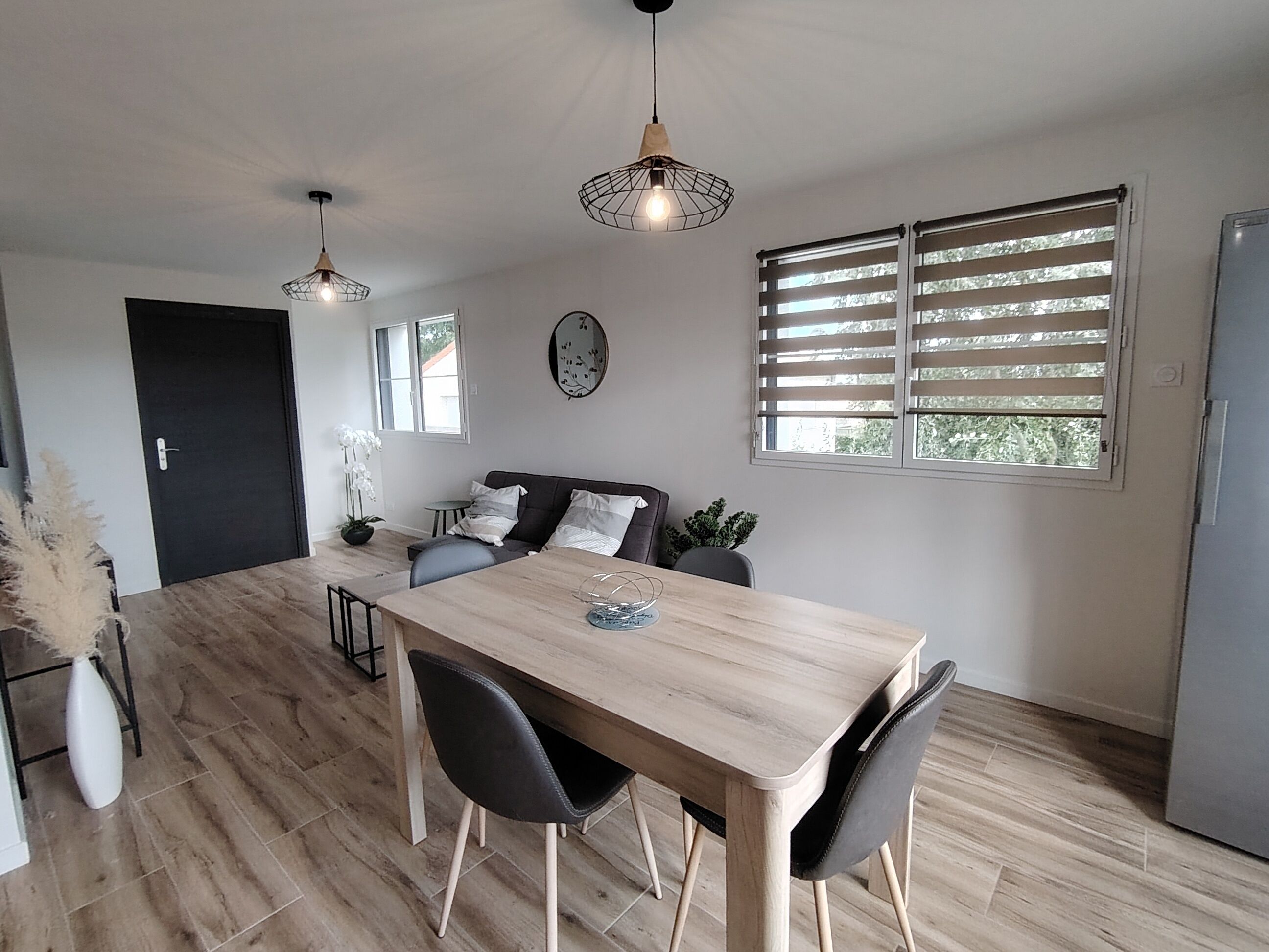 A modern kitchen with a wooden table, black chairs, pendant lights, and a window with blinds.