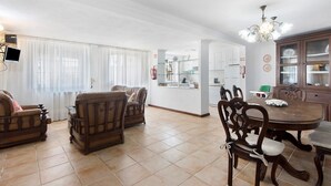 Ein Essbereich mit einem runden Tisch und Stühlen, im Hintergrund eine Küche und ein Wohnzimmer mit Sofas und einem Fernseher.