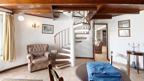 Ein Zimmer mit einer Wendeltreppe, einem gepolsterten Sessel, einem Esstisch mit blauer Tischdecke und einer Holzbalkendecke.