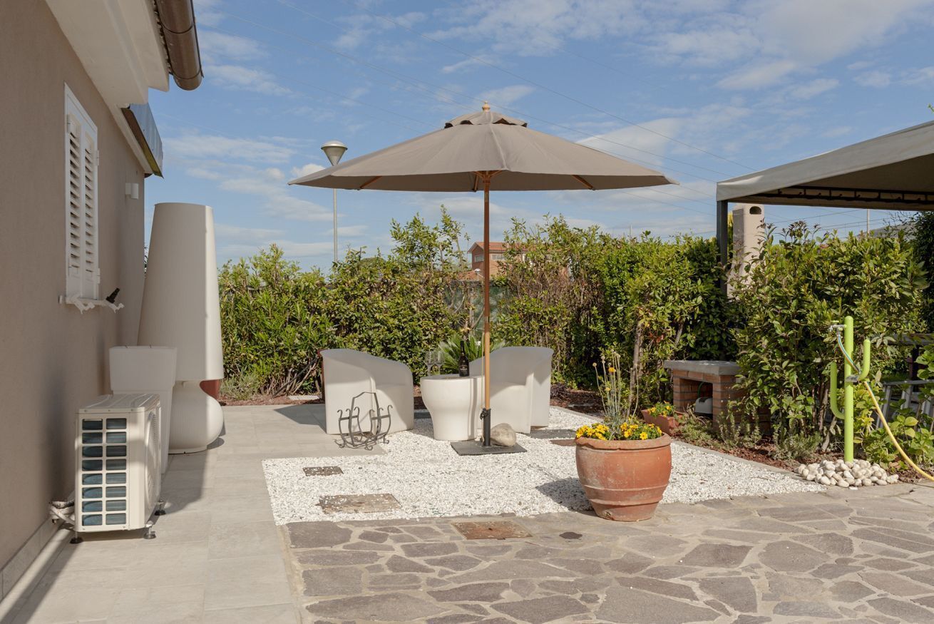Eine Terrasse im Freien mit einem großen Sonnenschirm, weißen Stühlen, einem Blumentopf und einem Steinfußboden.