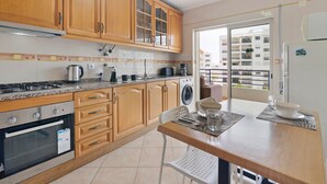 A kitchen with wooden cabinets, a gas stove, a washing machine, and a dining area with a table and chairs. - Apartment 'Rei  3º T' with Balcony, Wi-Fi and Air Conditioning (Quarteira)