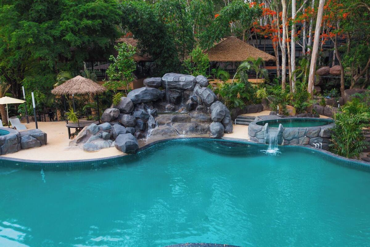 Una piscina con caída de agua rodeada de exuberante vegetación tropical y construcciones de techo de paja.