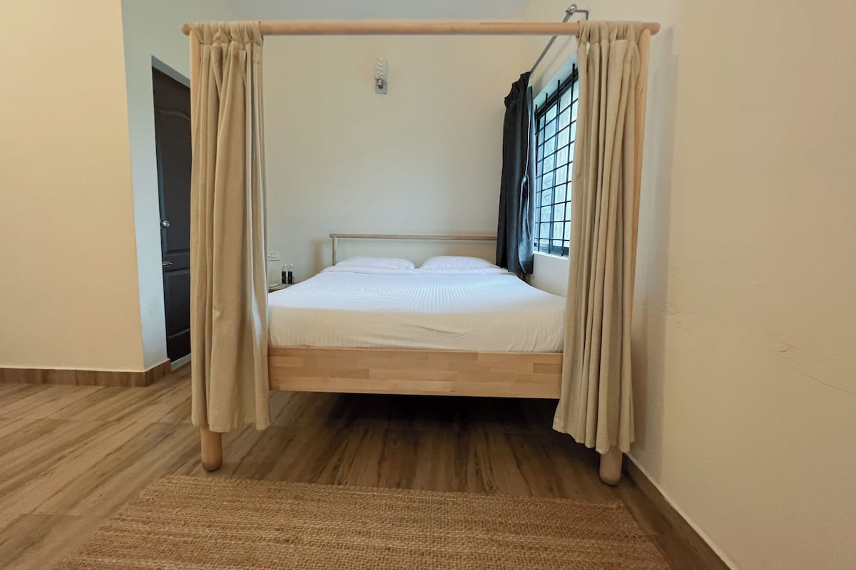 Una cama individual con estructura y cabecera de madera, una ventana con cortinas, una puerta y piso de madera.