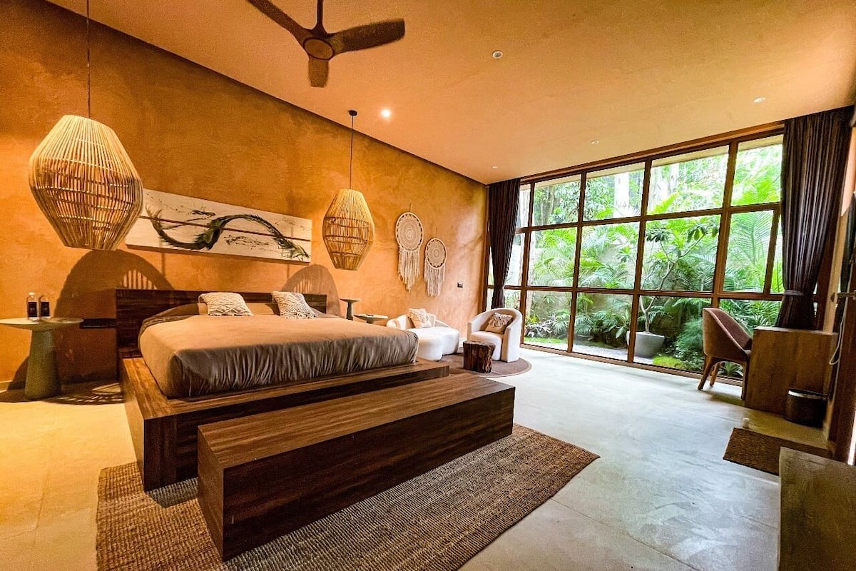 Un dormitorio moderno con una cama grande, mesitas de noche, un banco, un ventilador de techo y ventanas amplias con vistas a zonas verdes.