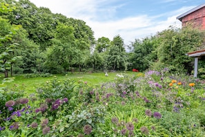 Um jardim exuberante com diversas plantas e flores, um gramado e um edifício de tijolos ao fundo.