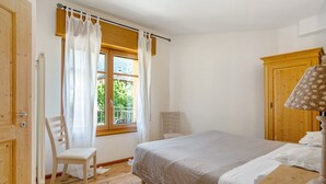Quarto com piso de madeira, uma cama com colcha branca, uma cadeira, um guarda-roupa de madeira e uma janela com cortinas.