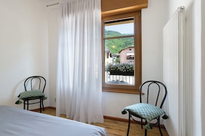 Quarto com duas cadeiras, uma cama, janela com vista para casas e vegetação, e um radiador.