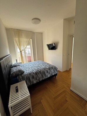 A bedroom with a bed, a TV mounted on the wall, a small table, and a window with curtains. - Modern three-bedroom apartment with a terrace and parking space (Milano)