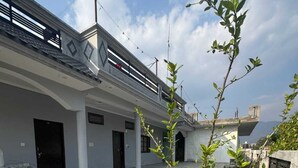 A rooftop terrace with potted plants, a red pipe, and a building with a balcony in the background.
