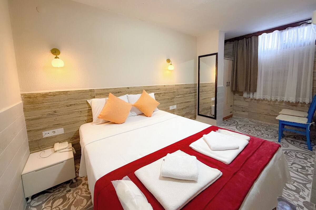 Habitación de hotel con cama, mesitas de noche, espejo, silla y ventana con cortinas.