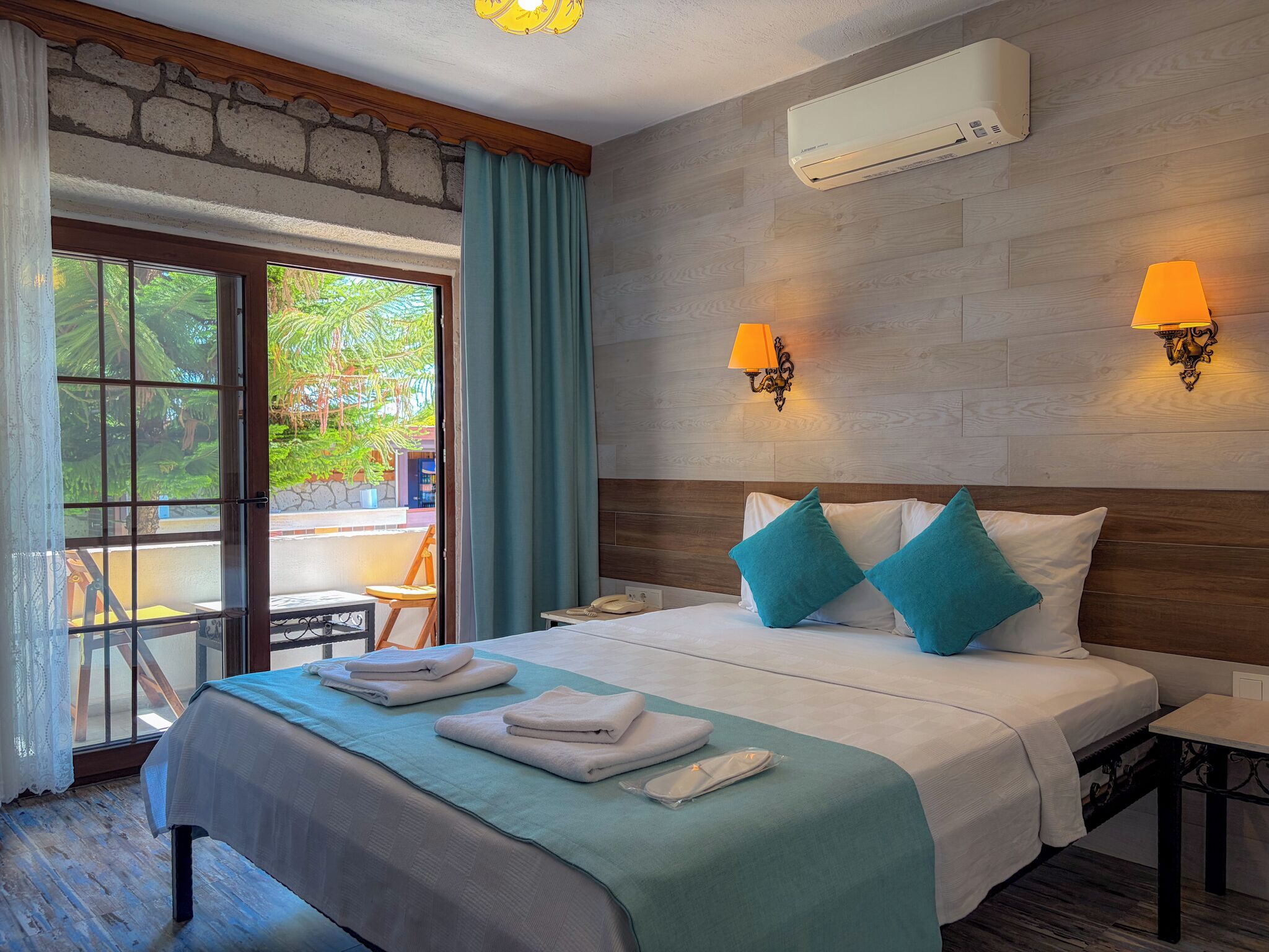 A hotel room with a bed, a nightstand, a table, a chair, an air conditioning unit, and a view of greenery outside.