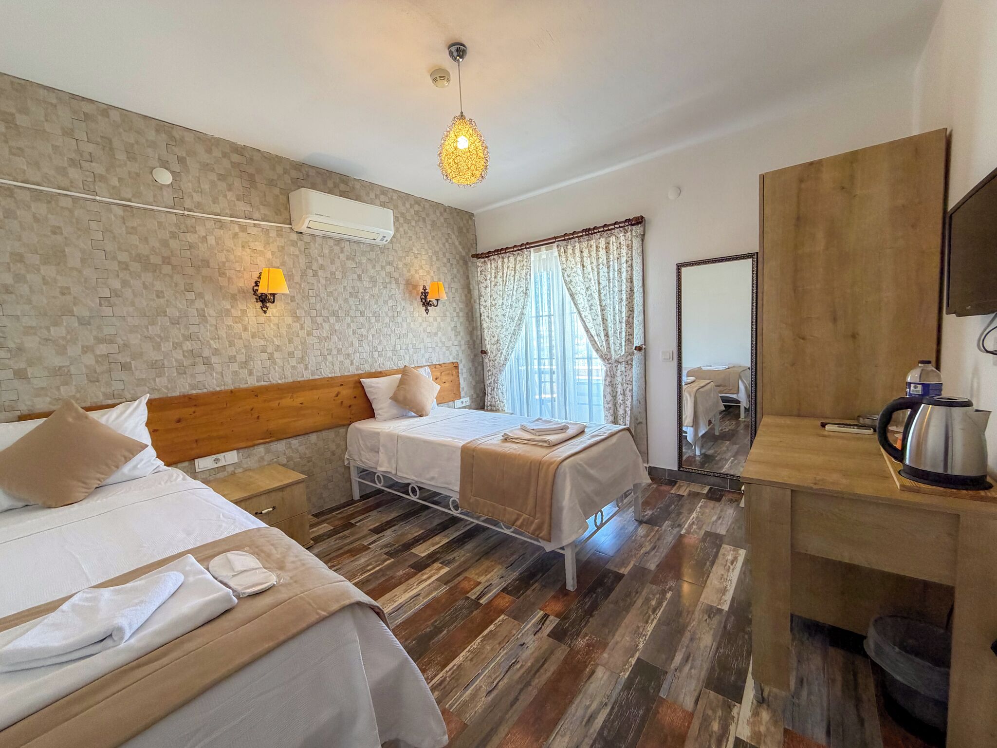 A hotel room with two beds, a wooden floor, a large mirror, a kettle, and a TV.