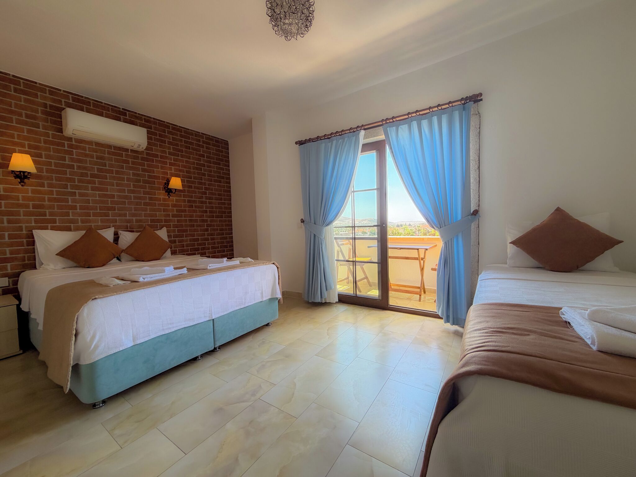 A hotel room with two beds, a brick wall, a balcony with a table and chairs, and a sliding glass door with blue curtains.