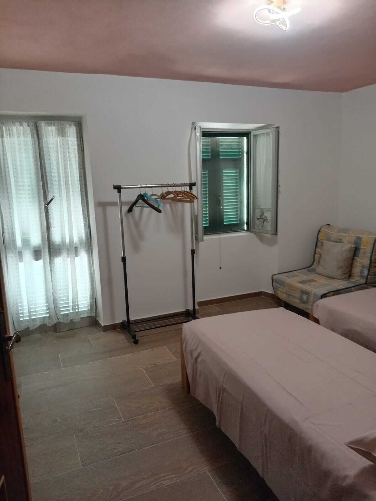 Ein Zimmer mit zwei Betten, einem Fenster mit Jalousien, einem Kleiderständer mit Kleiderbügeln und einer Tür mit Vorhängen.