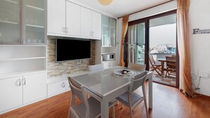 A modern kitchen with a flat-screen TV, white cabinets, and a dining area with a glass table and chairs. - Vivienne In Front Of Pool Holiday Home with Shared Pool, Wi-Fi & Air Conditioning (Desenzano del Garda)