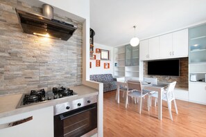A modern kitchen with a gas stove, oven, and a built-in range hood. There is a dining area with a table and chairs, and a TV mounted on the wall. - Vivienne In Front Of Pool Holiday Home with Shared Pool, Wi-Fi & Air Conditioning (Desenzano del Garda)