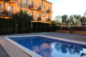 Une piscine rectangulaire avec un escalier en métal, entourée d’une terrasse carrelée. Au fond, on aperçoit un immeuble à plusieurs étages doté de balcons et bordé d’une haie.