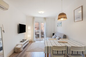 Een moderne, goed verlichte kamer met een televisie, een eettafel met stoelen, een bank en een balkon met tuinmeubilair.