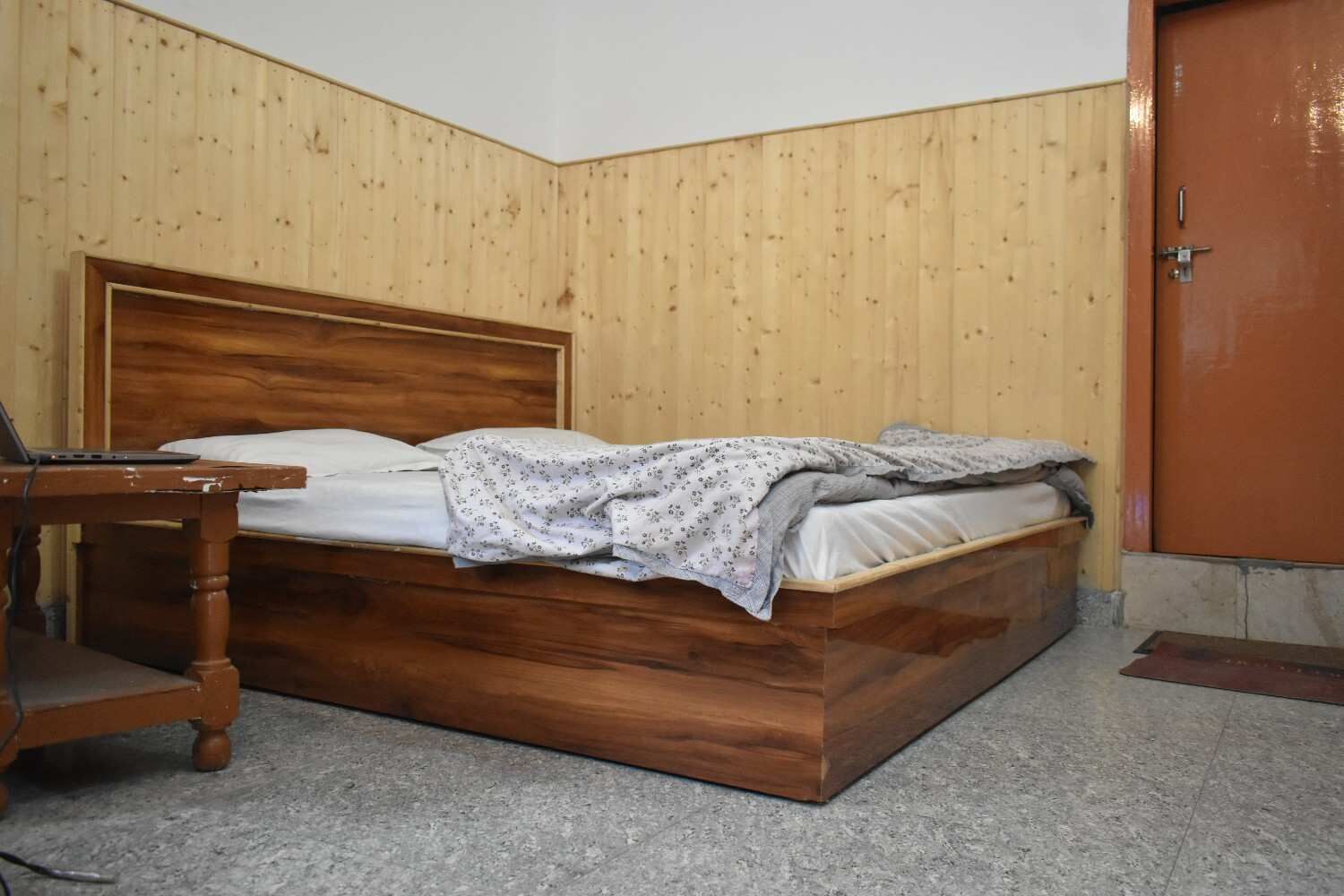 A single bed with a wooden headboard, a bedside table, and a door to the right.