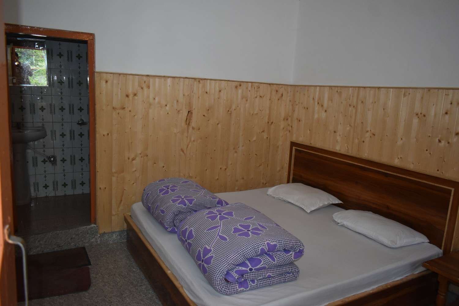A single bed with a wooden headboard, a bathroom with a tiled wall, and a wooden door.