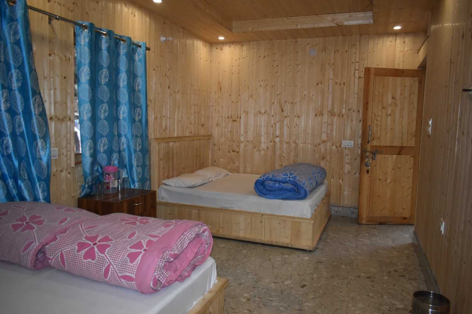 Una habitación con paneles de madera, dos camas, una puerta de madera y una pequeña mesa con una manta de diseño rosa y azul.