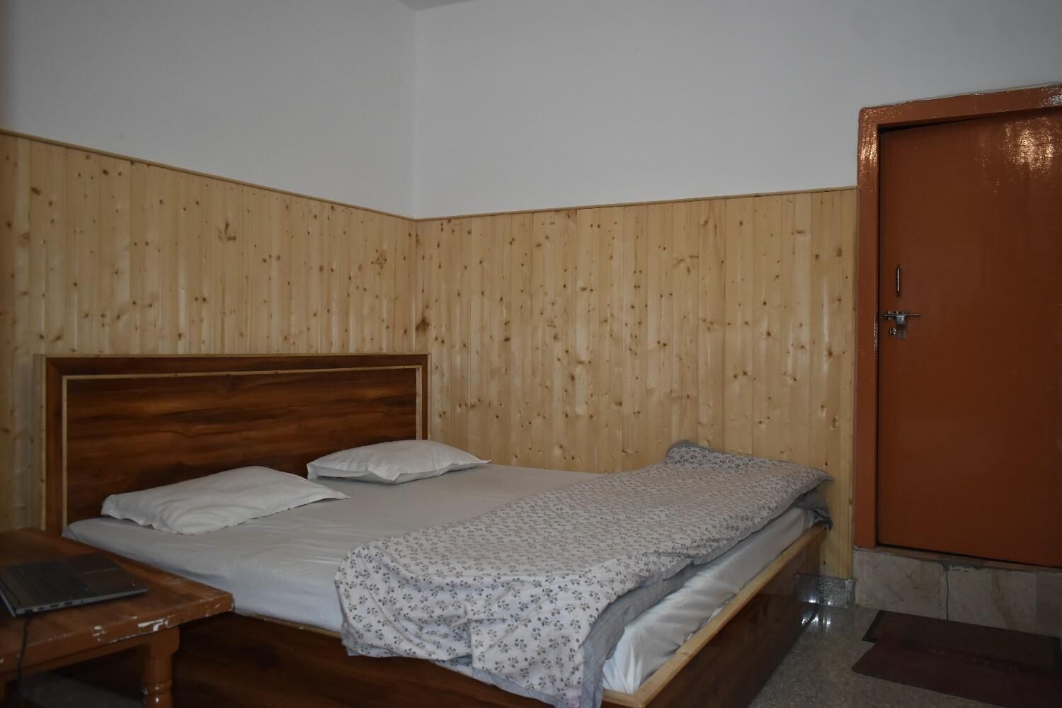 Una cama individual con cabecera de madera, una mesita de noche con una laptop y un armario de madera.