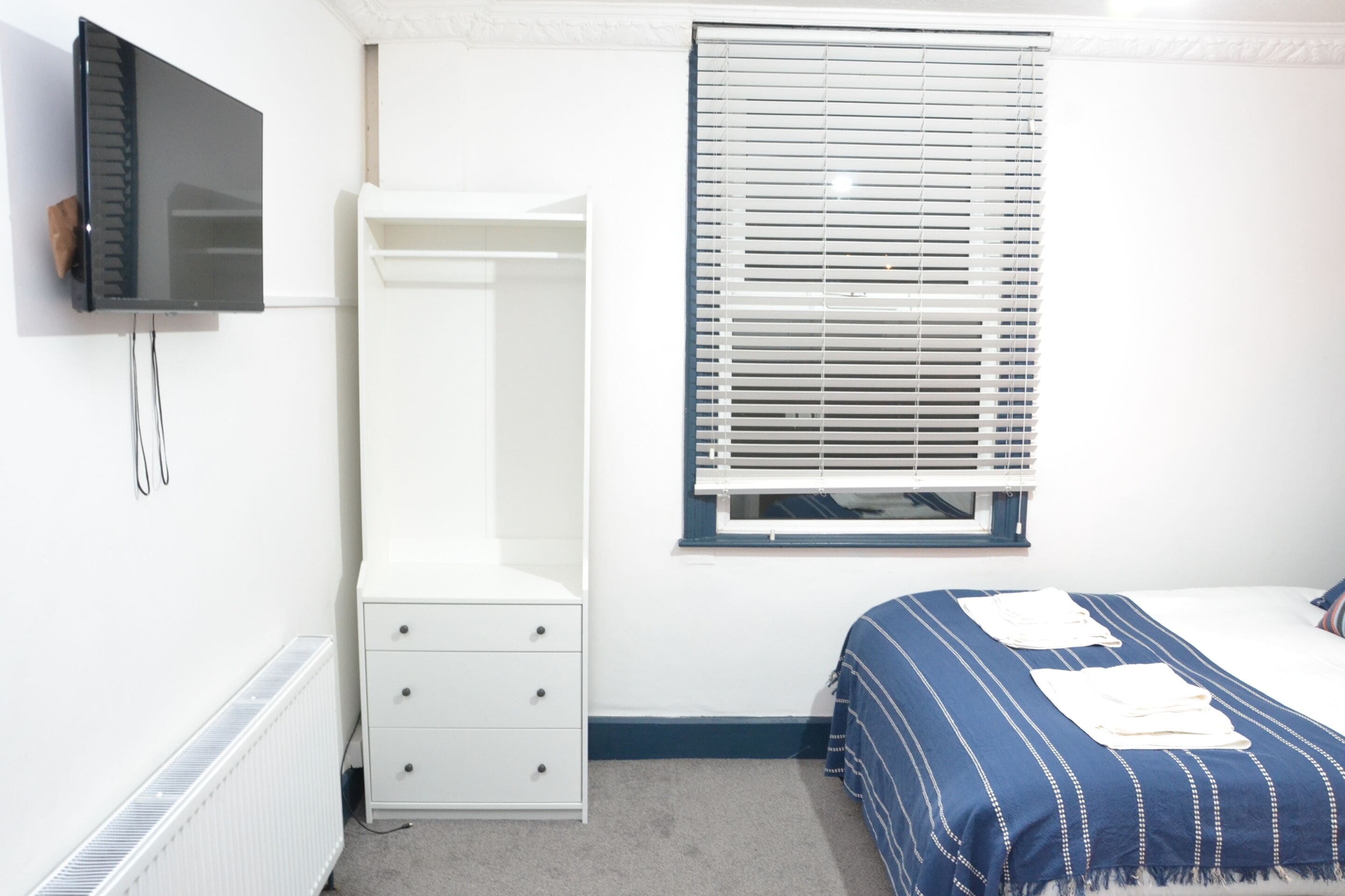 A bedroom with a bed, a white dresser, a TV mounted on the wall, and a window with blinds.